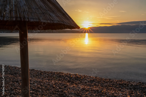 Fototapeta Naklejka Na Ścianę i Meble -  Straw umbrellas at sunset on idyllic pebble beach in tourist resort town Baska, Krk Otok, Primorje-Gorski Kotar, Croatia, Europe. Paradise summer atmosphere in Kvarner bay, Mediterranean Adriatic Sea