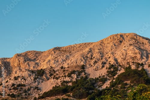 Panoramic view during sunrise of majestic mountains with massive rock formation in coastal town Baska, Krk Otok, Primorje-Gorski Kotar, Croatia, Europe. Illuminated mountain peak in soft red colours.