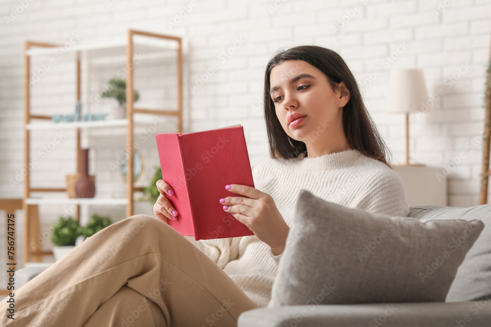 Beautiful young woman sitting on comfortable sofa and reading book in living room at home