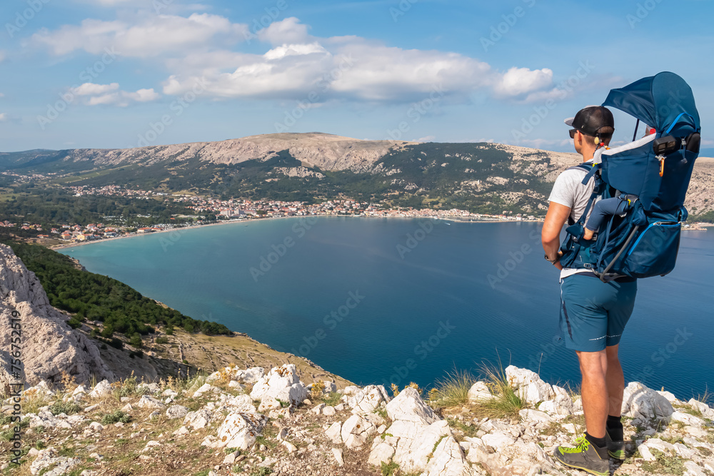 Naklejka premium Father with baby carrier on hiking trail with panoramic aerial view of idyllic lagoon in coastal town Baska, Krk Island, Primorje-Gorski Kotar, Croatia, Europe. Coastline of Mediterranean Adriatic Sea