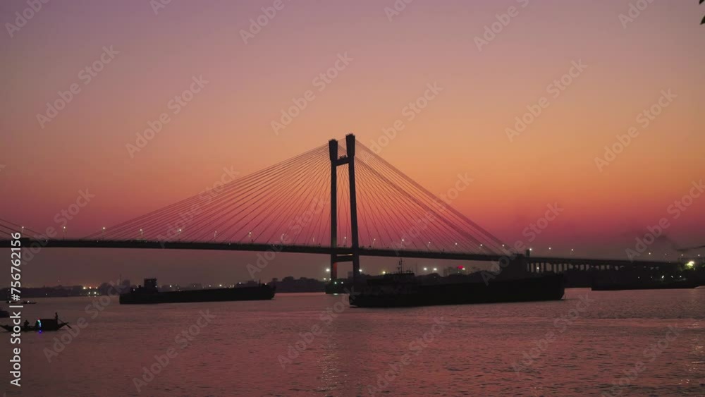 A silhouette wide shot of the modern cable-stayed Vidyasagar bridge against the vibrant gradient orange twilight sky after the sunrise with ships and vessels stationed in the backwater or river A silhouette wide shot of the modern cable-stayed Vidyasagar bridge against the vibrant gradient orange twilight sky after the sunrise with ships and vessels stationed in the backwater or river