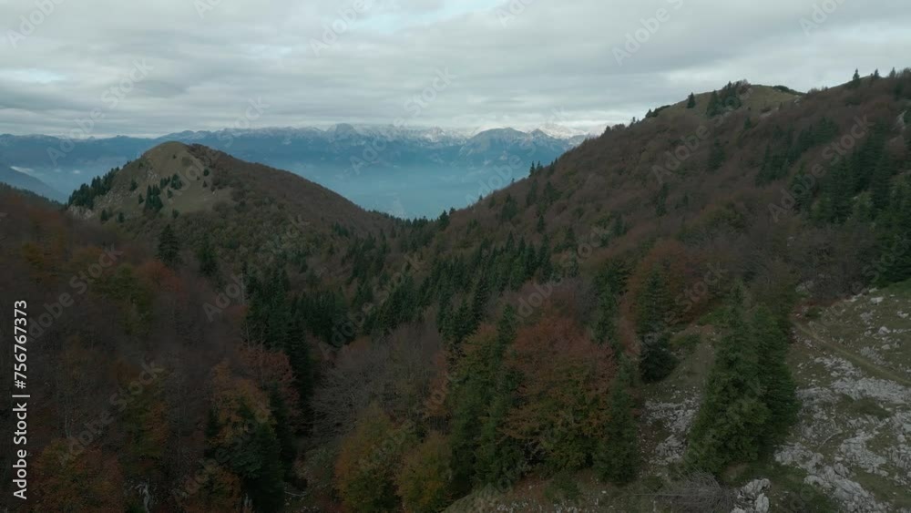 Drone shooting along wooded hills. In the background there are high mountains and a cloudy sky. Drone shooting along wooded hills. In the background there are high mountains and a cloudy sky.