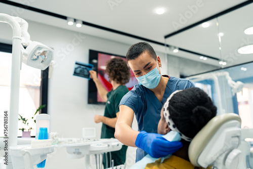 Female dental assistant preparing a patient for a dental surgery while the male oral surgeon is looking at a dental x-ray image at the modern dental clinic.