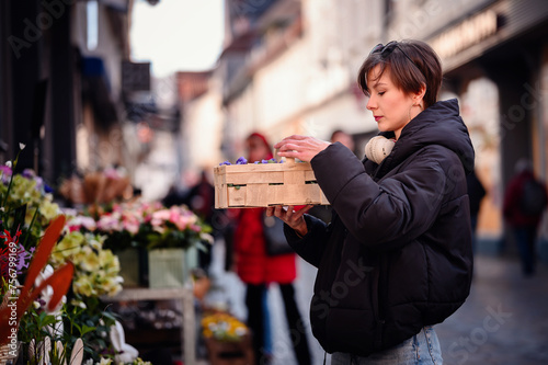 A stylish woman with headphones examines a crate of colorful flowers at a vibrant city market, showcasing a moment of everyday urban life