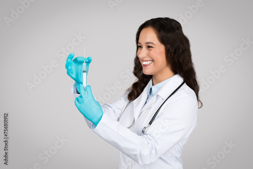 Cheerful young female doctor in white lab coat and gloves carefully preparing syringe for vaccination