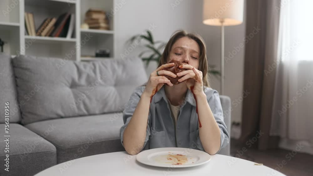 Portrait of funny happy young woman eating burger with great pleasure while ketchup flows down the hands at home Excited hungry female biting hamburger enjoying fast junk food indoors Portrait of funny happy young woman eating burger with great pleasure while ketchup flows down the hands at home Excited hungry female biting hamburger enjoying fast junk food indoors