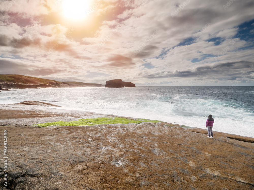 Teenager girl walking on stunning rough stone beach by the ocean on warm sunny day with cloudy sky. Kilkee area, Ireland. Popular tourist landmark with beautiful scenic Irish landscape.