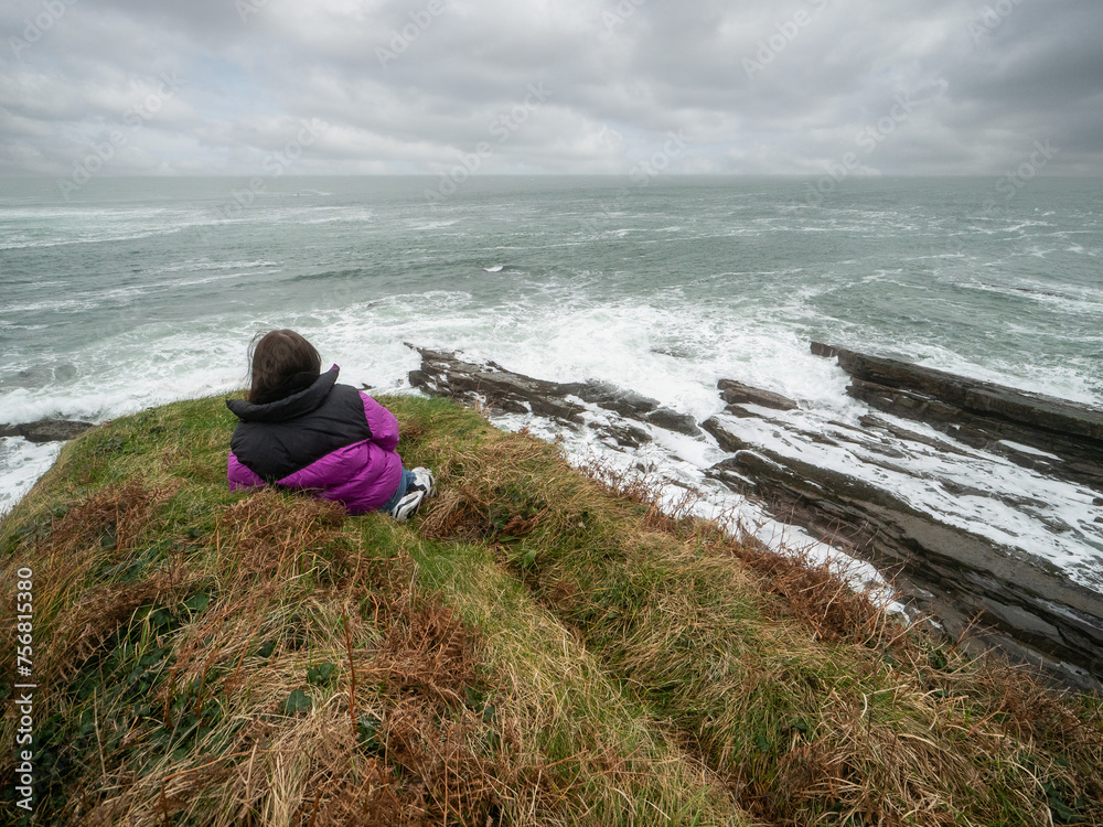 Teenager tourist girl laying on edge of a cliff and looking at stunning nature scene with rough stone coastline with cliff, ocean waves and dramatic sky. Mullaghmore, county Sligo, Ireland. Teenager tourist girl laying on edge of a cliff and looking at stunning nature scene with rough stone coastline with cliff, ocean waves and dramatic sky. Mullaghmore, county Sligo, Ireland.