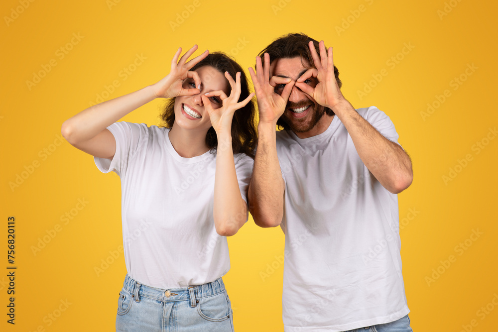 Playful couple making funny glasses with fingers over eyes, laughing in matching white t-shirts Playful couple making funny glasses with fingers over eyes, laughing in matching white t-shirts