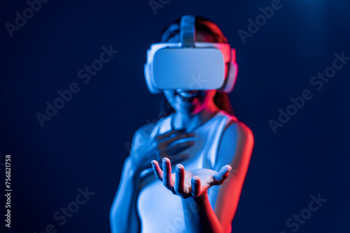 Fototapeta Naklejka Na Ścianę i Meble -  Smart Female standing surrounded by neon light wear VR headset connecting metaverse, futuristic cyberspace community technology. Elegant woman use hand holding generated virtual object. Hallucination.