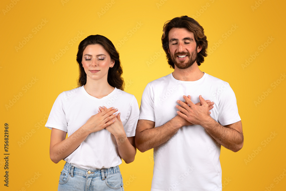 Grateful young couple with eyes closed and hands on heart, feeling thankful and serene in plain white t-shirts Grateful young couple with eyes closed and hands on heart, feeling thankful and serene in plain white t-shirts