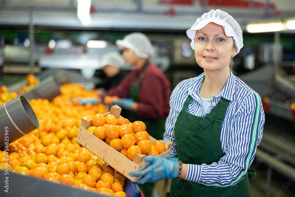 Positive female worker in fruit sorting and packing warehouse holding small wooden box of selected ripe mandarins
