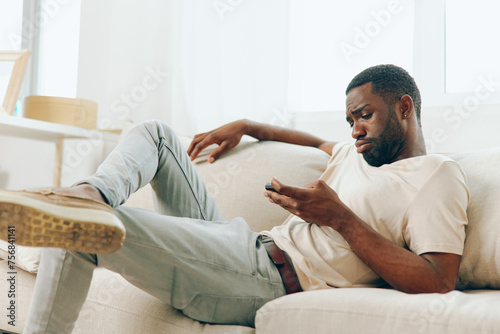 Happy African American Man Using a Black Smartphone while Relaxing on a Modern Sofa at Home