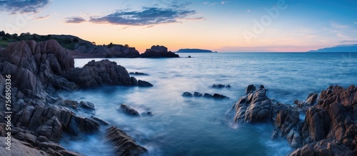 A breathtaking long exposure photo capturing the rocky shoreline at dusk, with the oceans waves crashing against the rocks under the colorful sky