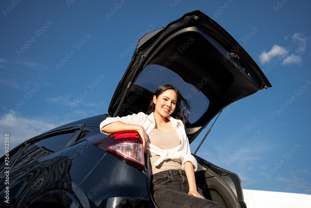 Young beautiful asian women buying new car. she was sitting at trunk of car. Beautiful moment blue sky Smiling female driving travel by vehicle on the road on a bright day weekend with sun light.