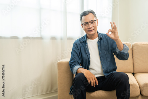 Fototapeta Naklejka Na Ścianę i Meble -  Handsome asian mature old man sitting on sofa couch in living room at home. Happy Portrait of cheerful smiling senior asian man relax in the house. Mature People and lifestyle