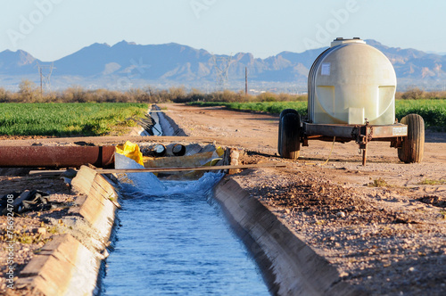 Fotografie Concrete irrigation canal delivers water to green crop fields, with a mobile tank trailer and distant desert mountains, illustrating agricultural water use and rural farm infrastructure