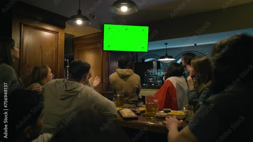 Group of multi ethnic friends sit in sports pub and watch live broadcast on TV Green Screen, chromakey. People gathered to cheer team playing in tournament. Crowd of fans support club during football