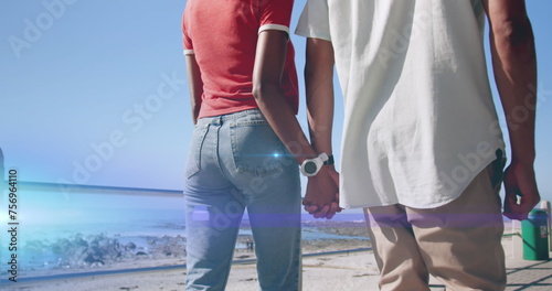 Image of light moving over midsection of couple walking by beach holding hands