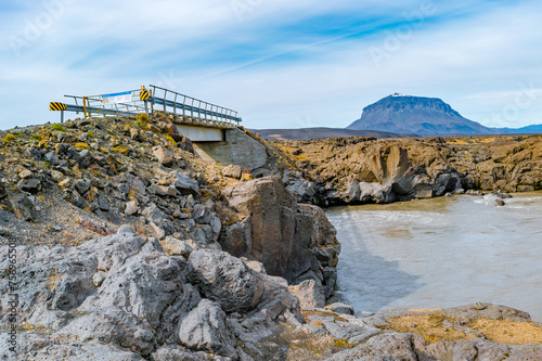 Glacial river, bridge Herdubreid volcanoe in the deadliest volcanic desert in Highlands, with stones and rocks thrown by volcanic eruptions, Iceland, summer, blue sky.