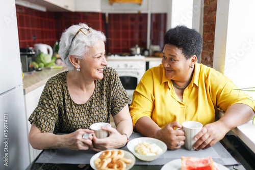 Two aged ladies of diverse ethnicity sitting at kitchen table drinking coffee, sharing gossips and rumors, discussing news, looking at each other with laughing faces, enjoying spending time together