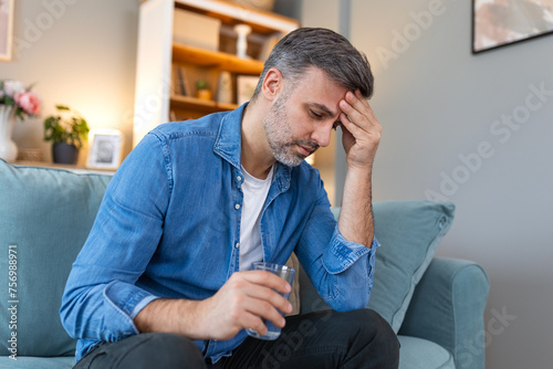 Man at home having a headache. Man suffering from headache after hard working day, sitting on couch at home holding glass of water