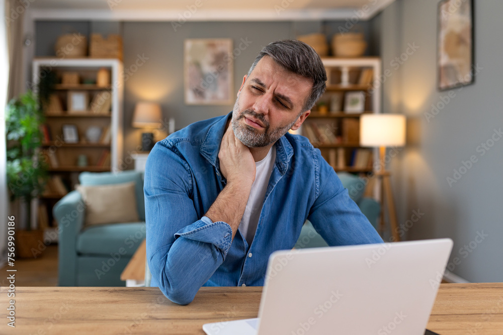 Obraz premium Businessman feeling pain in neck after sitting at the table with laptop. Tired man suffering of office syndrome because of long hours computer work. He is massaging his tense neck muscles