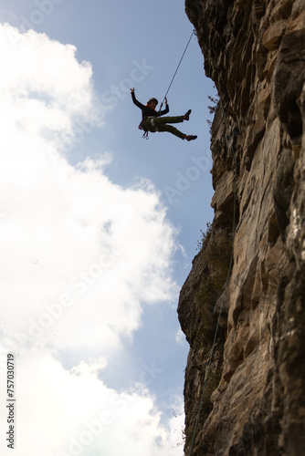 A man is hanging from a rock face with a rope. The sky is blue and there are clouds in the background