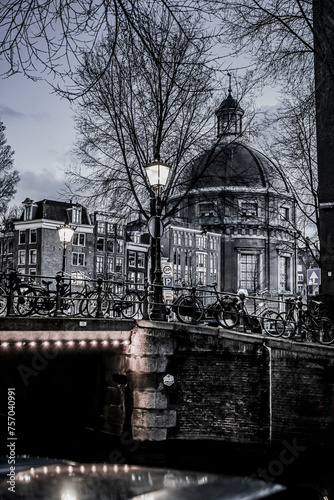 Evening view of a canal in Amsterdam featuring the Lutheran Church with its iconic dome. The dim streetlights and bare winter trees create a peaceful, atmospheric scene in the historic city