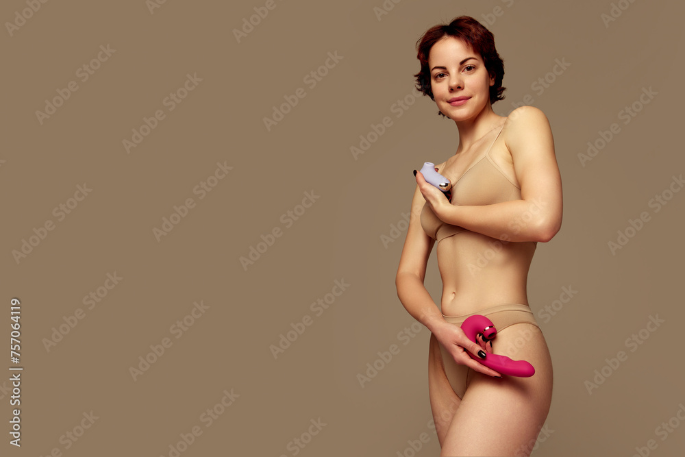 Smiling young girl in underwear standing with two vibrator toys against beige background. Concept of female health care, toys for adults, sexuality, pleasure. Banner. Empty space to insert text, ad Smiling young girl in underwear standing with two vibrator toys against beige background. Concept of female health care, toys for adults, sexuality, pleasure. Banner. Empty space to insert text, ad