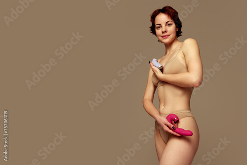 Smiling young girl in underwear standing with two vibrator toys against beige background. Concept of female health care, toys for adults, sexuality, pleasure. Banner. Empty space to insert text, ad