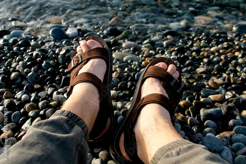 Bild auf Leinwand Man tourist in leather slippers resting relaxing on pebblestone sea beach with small waves in sunny summer weather, feet close-up