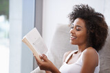 Happy, black woman and reading with book on couch for novel, story or literature in living room at home. African female person with smile for information, news or interesting knowledge at the house