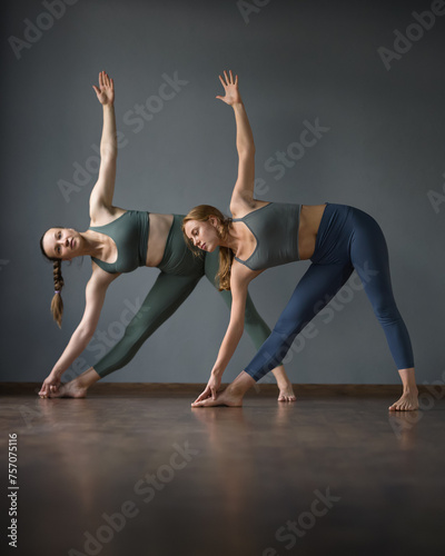 Two women doing yoga. Standing Triangle Pose. utthita trikonasana asana. training in Pilates, stretching in gym. Exercises to stretch leg. Wooden reflective surface. Gray background. Side view.