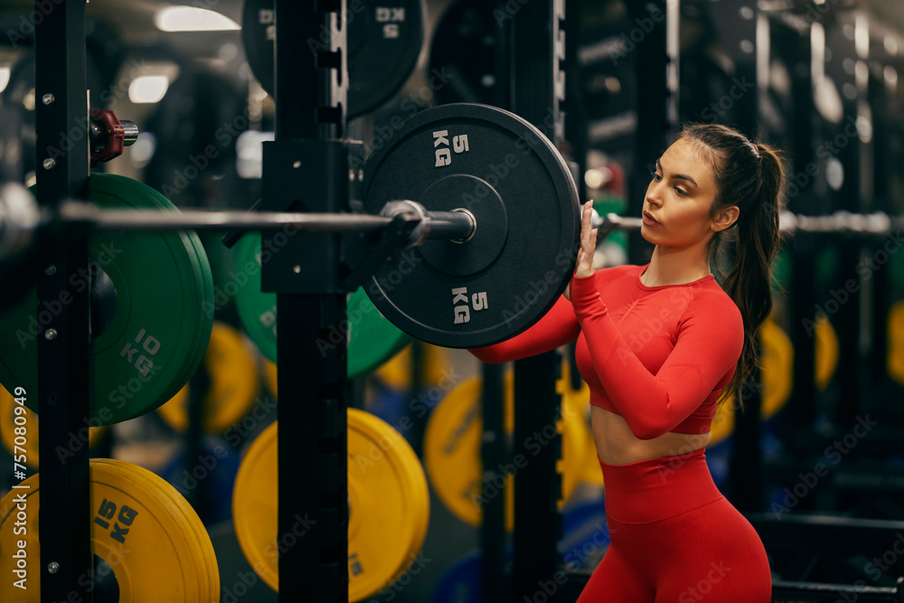 Portrait of a sportswoman putting weight plates on barbell in a gym. - obrazy, fototapety, plakaty 
