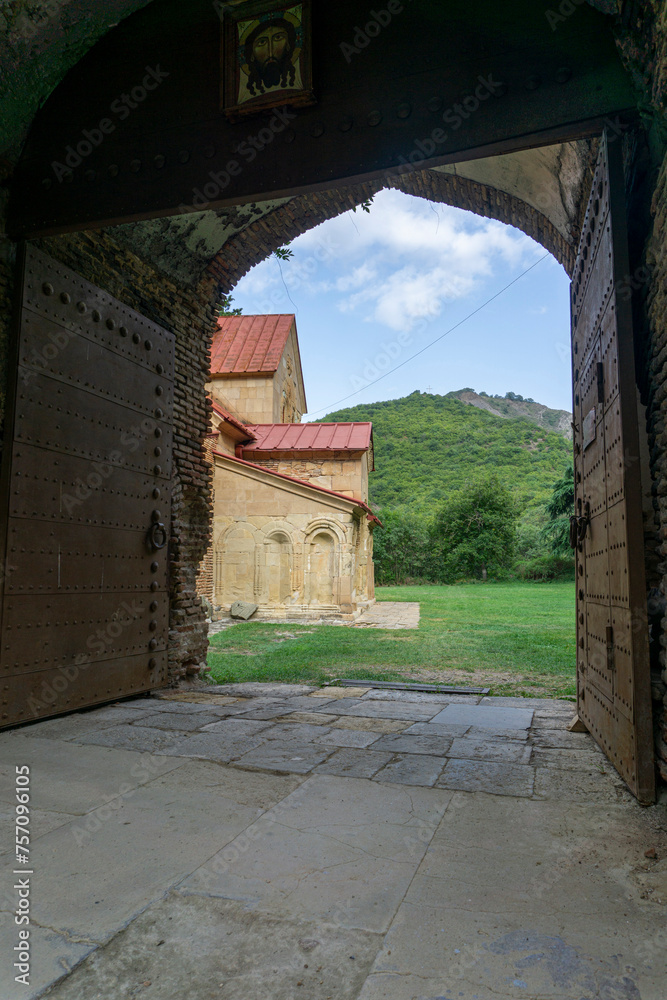 Fototapeta premium Church of the Nativity of the Mother of God of Betania monastery. View of the church through the open gate. Brick arch. Stone walls, iron roof. Bright blue sky.