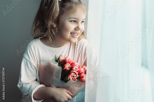 Beautiful little vaucasian girl looking at window empty copy space. Kid with flowers bouquet.Mother's day. Women's day.Birthday holiday.