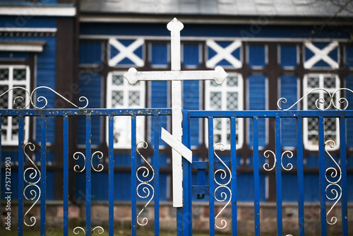 Orthodox cross isolated. Wooden church in the background. Blue paint metal faith symbol. East of Poland. Metal gate fence. Orthodox wooden church. Countryside small church made of blue wood.