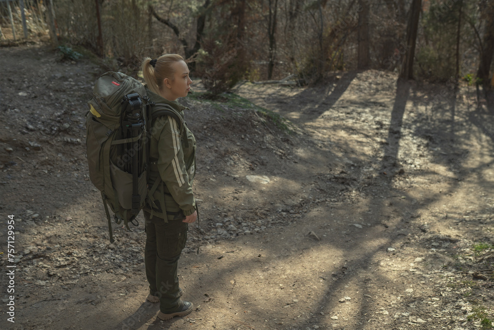 A hiker with a large backpack stands at a trailhead, looking onward into a dense forest. The sunlight breaks through the trees, casting long shadows on the path
