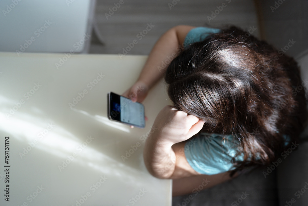 A pensive young girl sits at a table, deeply engrossed in the content on her smartphone. The indoor setting is bathed in natural light filtering through lace curtains, creating a tranquil domestic sce A pensive young girl sits at a table, deeply engrossed in the content on her smartphone. The indoor setting is bathed in natural light filtering through lace curtains, creating a tranquil domestic sce