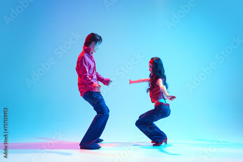 Happy, positive young man and woman in retro clothes dancing boogie woogie retro dance against blue background in neon light. Concept of hobby, dance class, party, 50s, 60s culture, youth photo