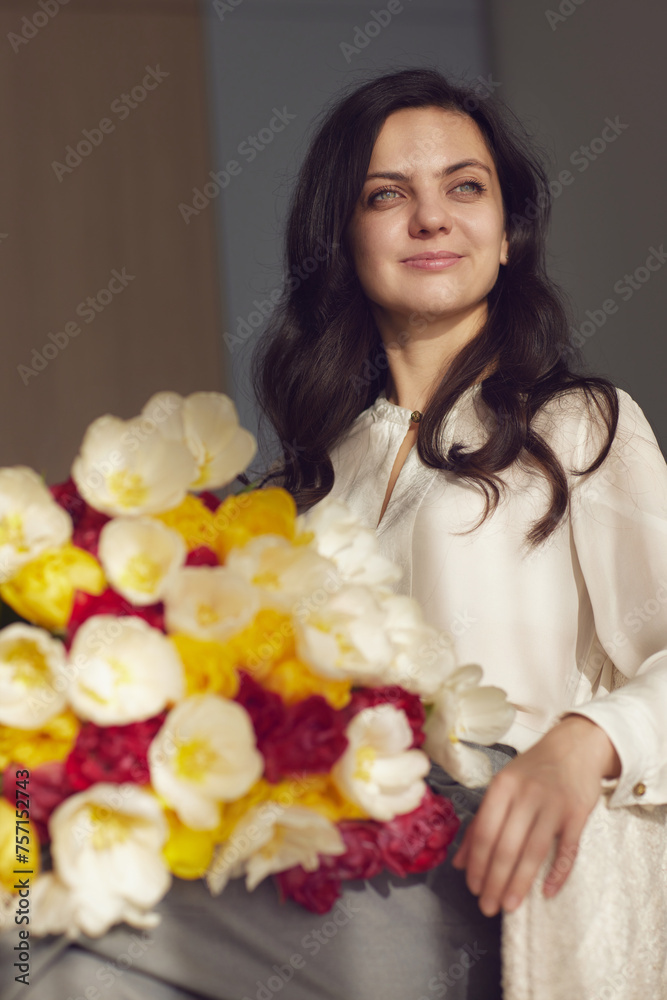 Beautiful woman with spring flowers tulips in hands sitting on chair near window. Women's Day Beautiful woman with spring flowers tulips in hands sitting on chair near window. Women's Day