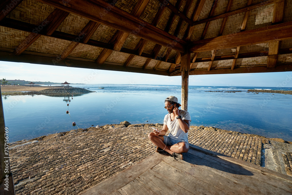 Relaxed and cheerful. Listening to music. Outdoor portrait of happy young african man resting on deck near the sea.