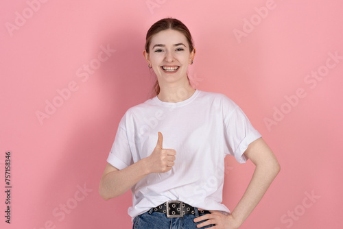 Attractive Caucasian young brunette woman in casual white t-shirt looks at the camera, smiles and shows thumb up isolated on pink studio background.
