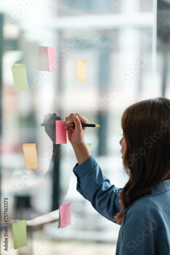 Beautiful female employee write down on colorful sticky notes manage list, concentrated biracial woman work on startup brainstorm collaborate plan on stickers on glass wall.