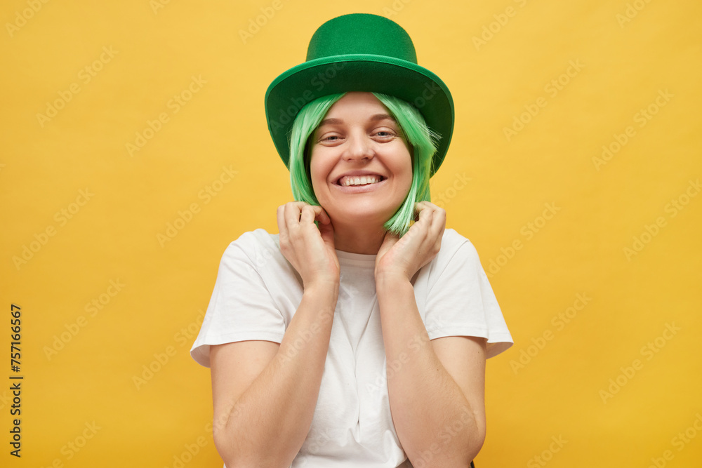 Fun festivities on St. Patrick's Day. Cute charming woman with green hair wearing leprechaun hat standing isolated over yellow background looking at camera with toothy smile