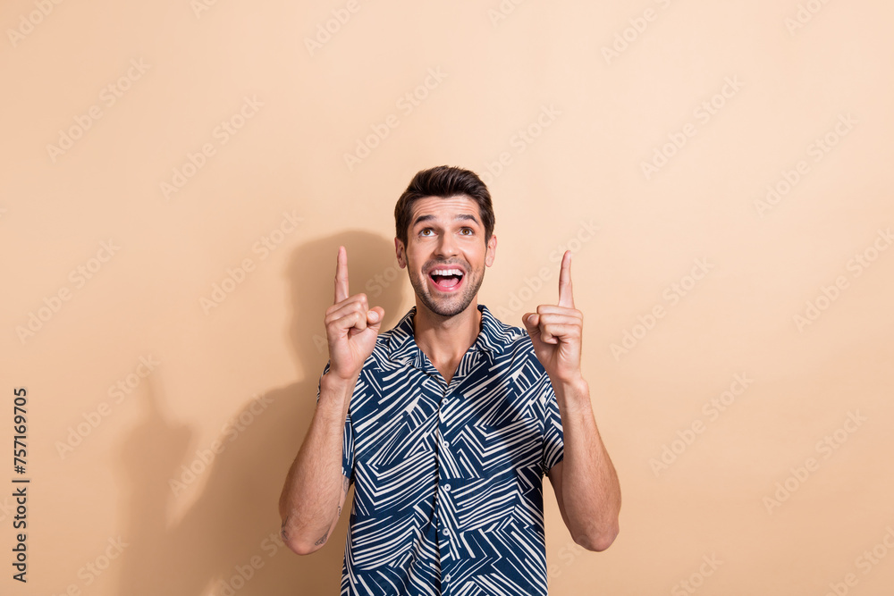 Portrait of overjoyed guy with trendy hairdo wear stylish shirt indicatign look up at sale empty space isolated on beige color background