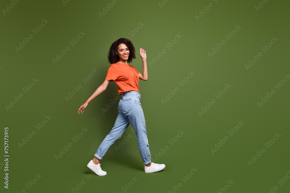 Full length photo of adorable friendly woman wear orange t-shirt waving arm hi walking empty space isolated green color background Full length photo of adorable friendly woman wear orange t-shirt waving arm hi walking empty space isolated green color background