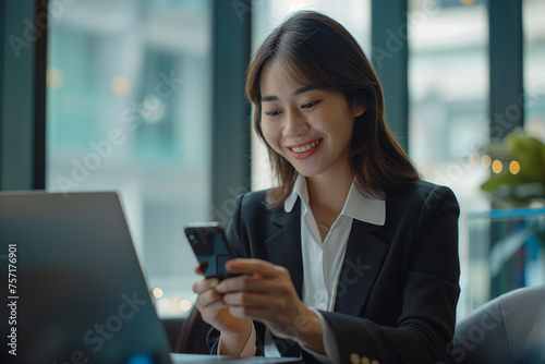 Young smiling asian businesswoman using and looking at mobile phone during working on laptop computer at modern office. Business woman in business suit online working and using smartphone photo