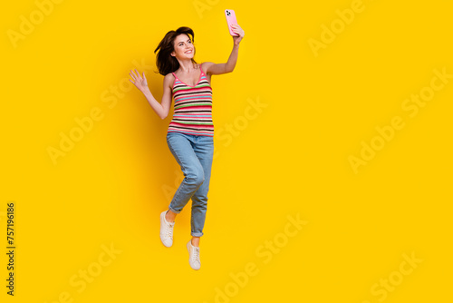 Full size photo of girlish woman dressed knit top jeans jumping making selfie on smartphone isolated on bright yellow color background